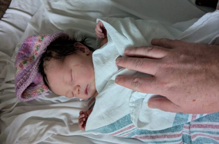 A newborn baby with dark hair wears a knitted hat and is wrapped in a blanket, lying on a hospital bed. An adult hand gently rests on the baby, showing support and care in this tender moment.