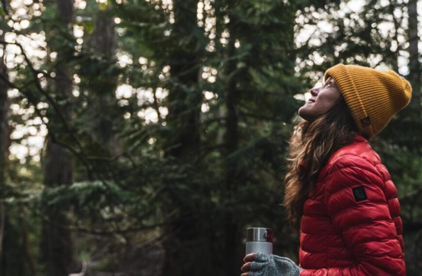 A person in a red jacket and yellow beanie holds a metal cup while looking up in a forest surrounded by tall trees, reflecting on how corporate philanthropy can double your donation to support nature conservation.