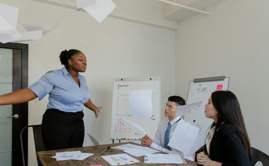 A woman stands and gestures while papers fly in the air, as two seated colleagues look on, highlighting the importance of emotional intelligence in managing high-pressure meeting room scenarios with charts and documents present.
