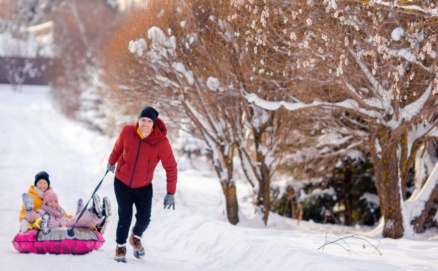 An adult pulls two children on a sled along a snowy path lined with trees, where the crisp winter air reminds us to stay vigilant about Lyme disease diagnosis and treatment, even during colder months.