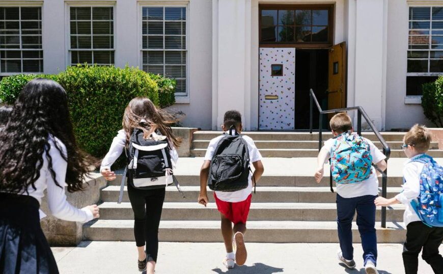 Five children with backpacks walk up the steps toward the entrance of a school building on a sunny day, ready for another exciting day of education in 2025.