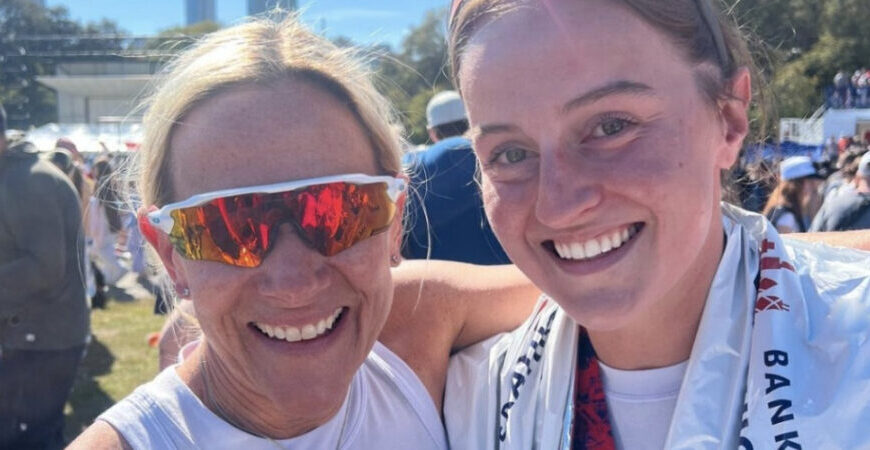 Two women in athletic attire smile for a photo outdoors at a crowded event, celebrating after one of the month of marathons. One wears reflective sunglasses; both are happy and sporting race medals or foil blankets.
