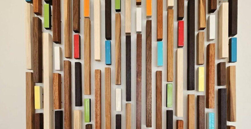 Vertical wooden blocks of varying lengths and colors, including natural wood tones and painted sections, are arranged in a geometric, wall-mounted pattern—an original artwork ideal for clinics seeking a modern touch.