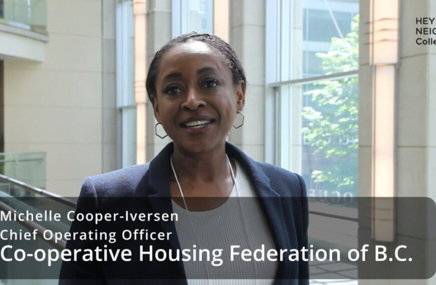 A woman in business attire stands indoors beside large windows. A text overlay introduces her as Michelle Cooper-Iversen, COO of Co-operative Housing Federation of B.C., during an interview.