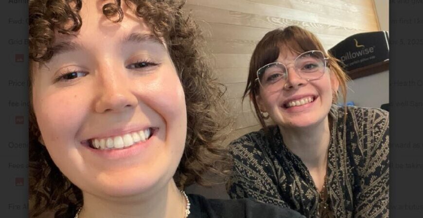 Two young women with curly and straight hair, smiling at the camera indoors like superheroes, one wearing glasses and a patterned shirt.
