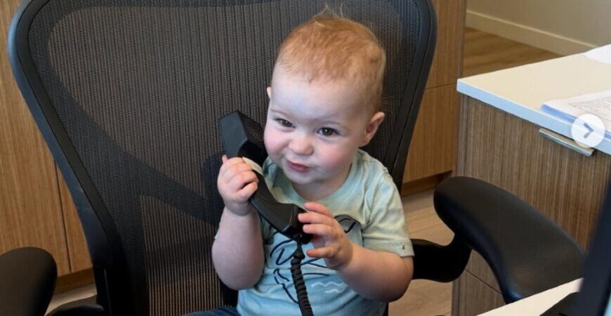 A baby sits in an office chair holding a telephone receiver to their ear, appearing to listen intently—like the youngest team member ready to take an important call.