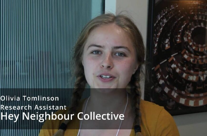 A young woman with braided hair stands indoors, wearing a yellow top and name tag. Text overlay reads: “Olivia Tomlinson, Research Assistant, Hey Neighbour Collective,” during an interview.