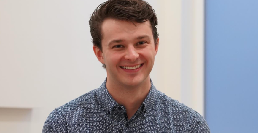 Josh, a young man with dark hair wearing a blue patterned button-up shirt, smiles while sitting indoors against a light background at Westshore physio.