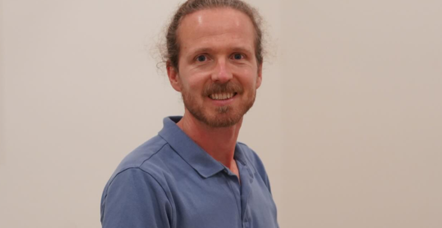 Remy, with light skin and brown hair tied back, wearing a blue polo shirt, stands indoors against a plain light-colored wall and smiles at the camera—ready to help as your Westshore physio.