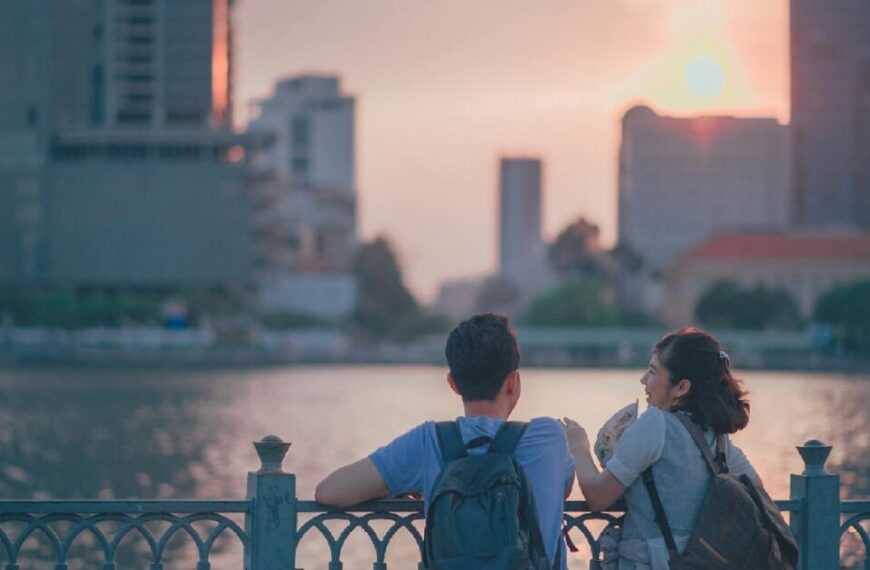 Two people with backpacks stand by a riverside railing, discussing the new term "underconsumption core" against a city skyline and sunset, reflecting on shifts in consumer culture.
