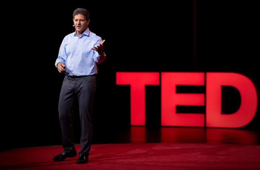 A man in a blue shirt and gray pants speaks on stage with large red "TED" letters in the background, exploring how the laws of economics shape our world.