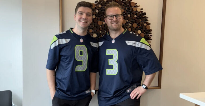 Two people stand indoors wearing matching navy blue Seahawks jerseys with the numbers 9 and 3, smiling at the camera.