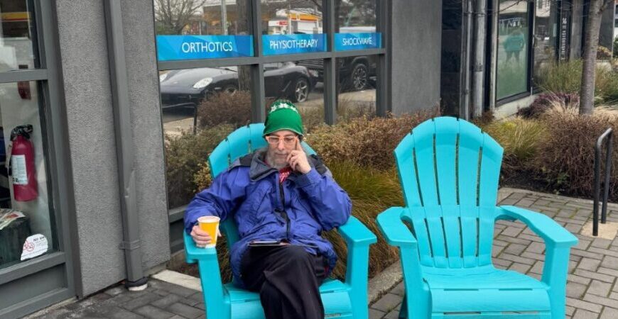 An older man in a blue coat and green hat sits on a turquoise outdoor chair outside a clinic, holding a coffee cup and looking at the camera. An empty chair is beside him, suggesting outdoor chairs are available for relaxation or even chairs for sale.