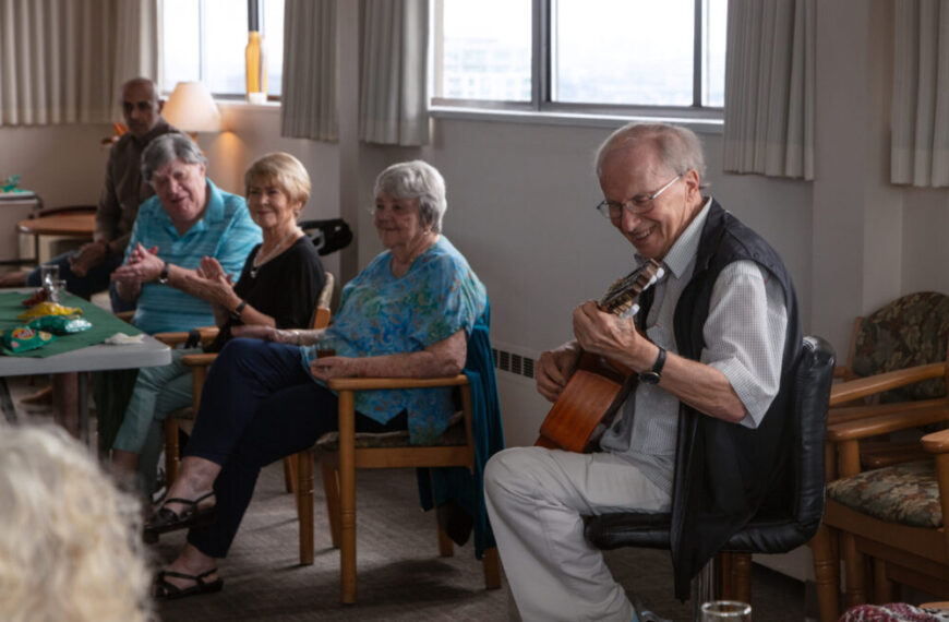 An elderly man plays guitar for a small group of seniors in a bright room, fostering neighbourly connections as some clap and smile, enjoying the moment together.