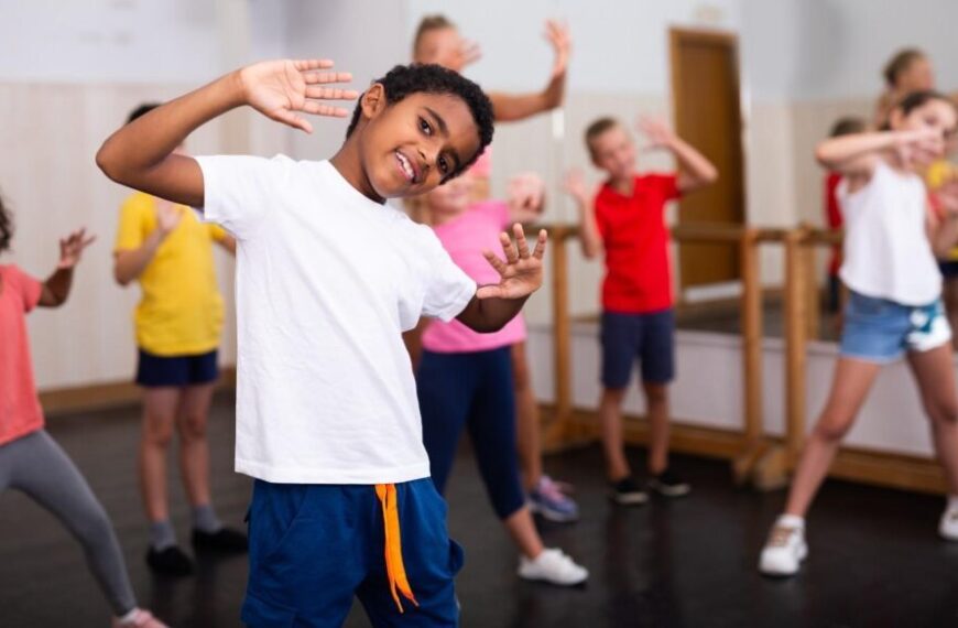 Children in casual clothes participate in a dance class, following movements together in a studio with mirrors and a wooden floor, embracing the principles of somatic therapy to encourage physical healing through movement.
