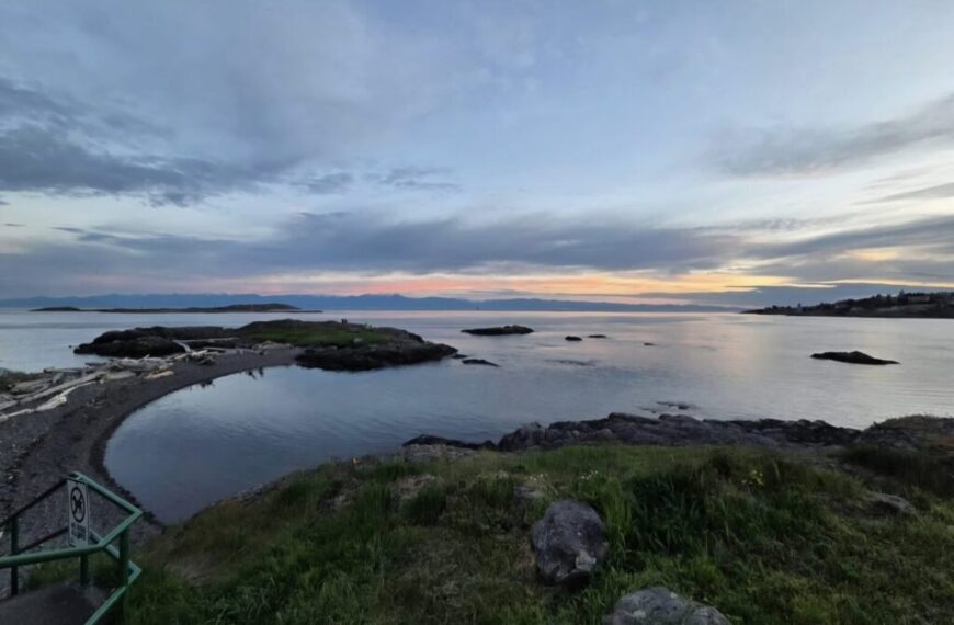 Rocky shoreline and grassy area overlooking calm water at sunset, with scattered clouds and distant land visible on the horizon—a peaceful scene ideal for attuning to the rhythms of the Fall Season.