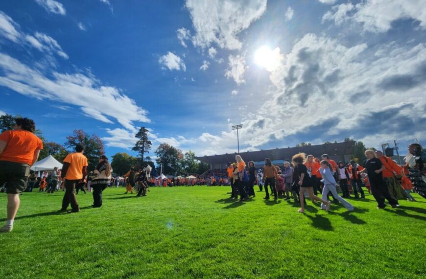 People gather on a sunny, grassy field, some wearing orange shirts to mark the National Day of Truth and Reconciliation on September 30. The sky is partly cloudy with the sun shining brightly overhead, while tents and trees are visible in the background.