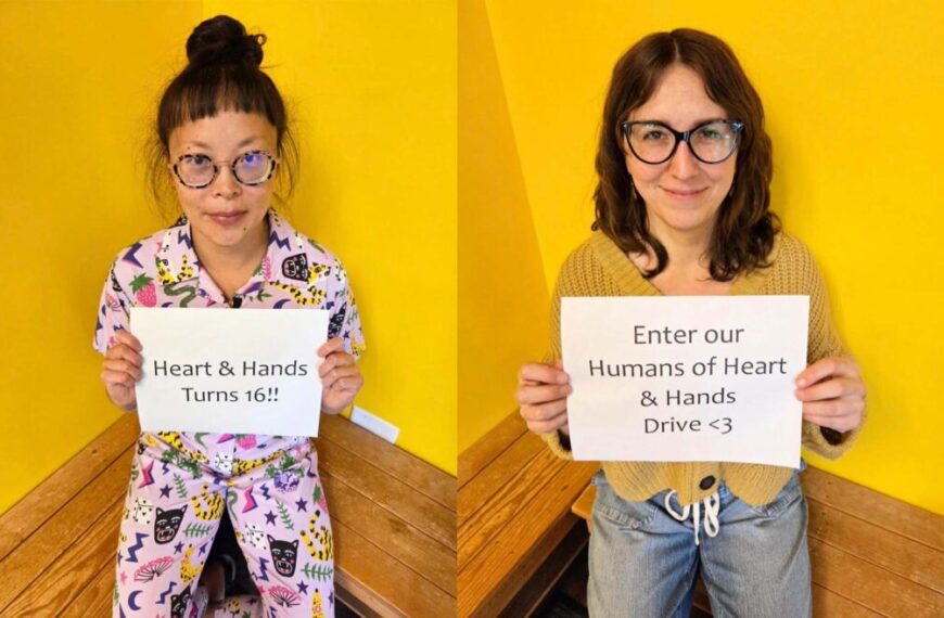 Two people sitting against a yellow wall each hold signs; one reads “Heart & Hands Turns 16!!” and the other reads “Enter our Humans of Heart & Hands Drive <3” in celebration of the H&H anniversary and upcoming donation drive.