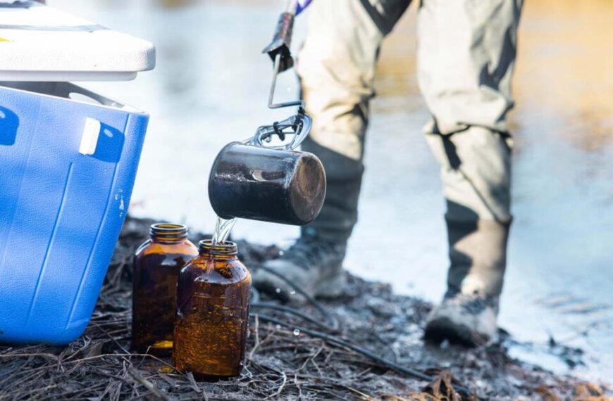 Person in waders pours water from a black container into brown glass bottles beside a blue cooler near the Fraser River Estuary, likely collecting samples to monitor hydrocarbon levels that could impact local salmon.
