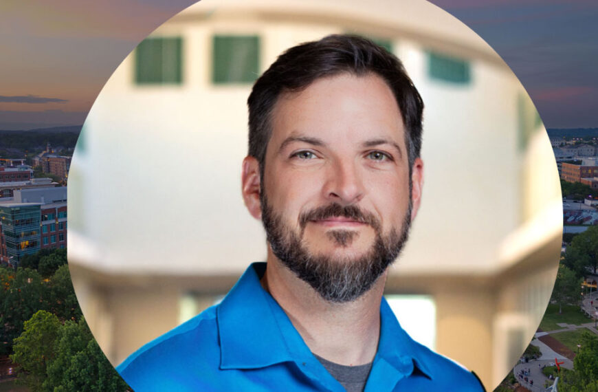 A man with short dark hair and a beard, wearing a blue shirt, stands indoors with a blurred background that adds depth to the scene.