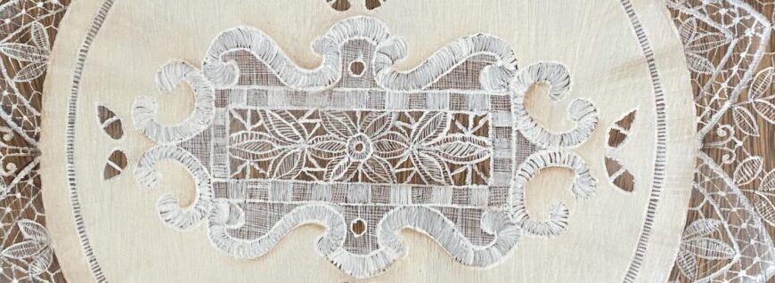 Close-up of an ornate white lace doily with floral and leaf patterns, placed on a wooden surface, as if discovered at a charming flea market or draped elegantly over the back of a vintage chair.