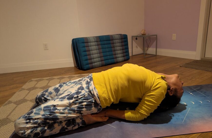 A Shiatsu practitioner is practicing fish pose on a yoga mat in a room with a wooden floor, a folded blanket against the wall, and a small table in the corner.