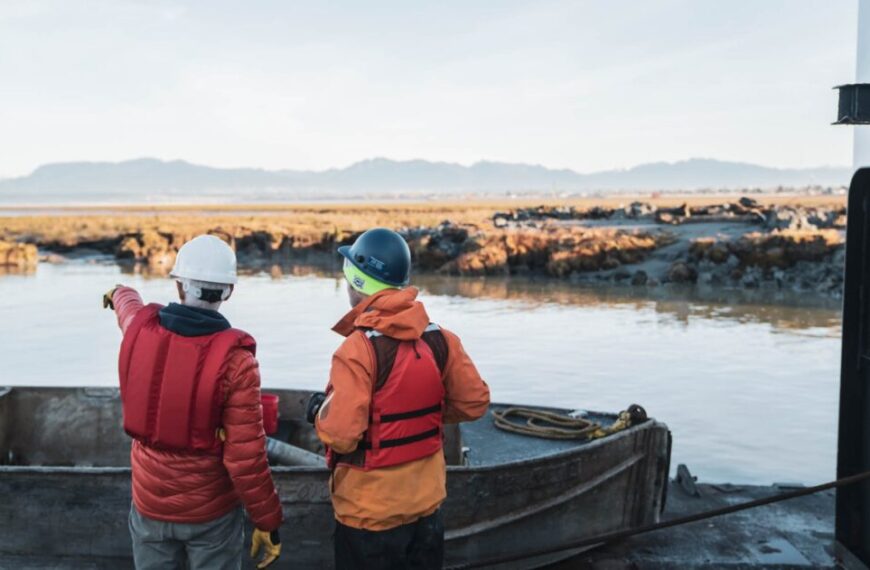 Two people in safety gear stand on a boat by the shore, discussing conservation finance as one points toward the water with distant mountains visible in the background.