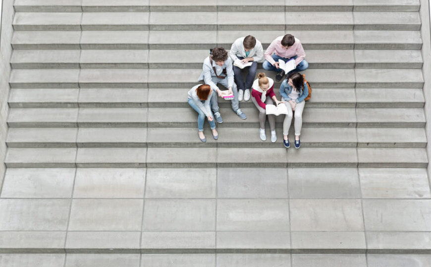 Five students sit on concrete stairs reading books while cats lounge nearby.